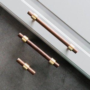 May include: Three wooden cabinet pulls with gold metal accents. The pulls are different sizes and are arranged in a staggered pattern on a white surface.