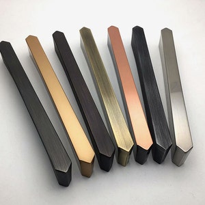 Könnte beinhalten: Acht Schrankgriffe in verschiedenen Ausführungen, darunter Schwarz, Gold, Kupfer und Silber. Die Griffe haben alle die gleiche Form, mit einem leicht abgewinkelten, rechteckigen Design.