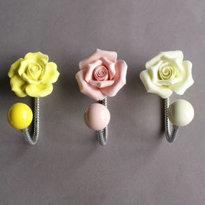 May include: Three decorative wall hooks with ceramic rose designs in yellow, pink, and off-white. Each hook has a matching spherical knob and a silver-toned, curved metal hook. The background is a solid grey.