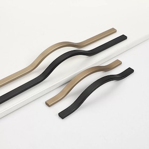 Può includere: Tre maniglie per armadi in diverse finiture. Due sono nere e una è dorata. Le maniglie sono curve e hanno un design moderno.