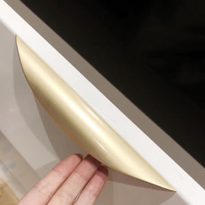 May include: A gold-coloured cabinet pull with a curved shape. The pull is mounted on a white cabinet door.