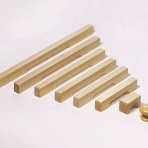 Może przedstawiać: Zestaw ośmiu drewnianych uchwytów do szafek w różnych rozmiarach, o prostym, prostokątnym designie i wpuszczanym uchwycie. Uchwyty są wykonane z jasnego drewna i mają gładkie wykończenie. Jest też mały, okrągły drewniany gałka.