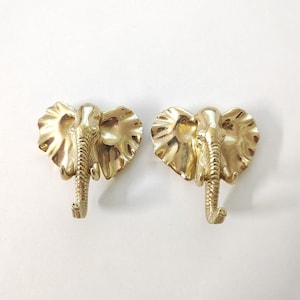 Pode incluir: Dois ganchos de cabeça de elefante de cor dourada. Os ganchos estão virados para a frente com as trombas enroladas.