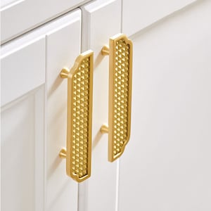 以下が含まれることがあります： 幾何学模様の金色のキャビネットプルが2つ。プルは白いキャビネットドアに取り付けられています。