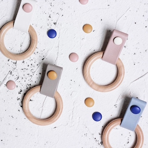 Op de afbeelding: Vier houten ringen met leren banden en gekleurde knopen. De ringen zijn in verschillende kleuren, waaronder wit, grijs, roze en blauw. De knopen zijn in tinten roze, geel en blauw.