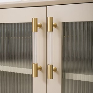 Peut inclure: Deux poignées de porte de placard dorées avec des inserts en verre transparent. Les poignées sont montées sur une porte de placard beige clair avec un panneau en verre dépoli.