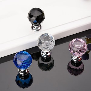 Pode incluir: Quatro botões de cristal para armário em diferentes cores: preto, transparente, rosa e azul. Cada botão tem um design facetado e uma base prateada.