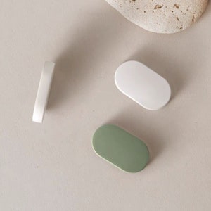 Pode incluir: Três puxadores de gaveta de cerâmica em forma oval. Um é branco, outro é verde e outro é branco com uma borda arredondada.