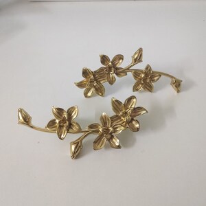 Puede incluir: Dos adornos de rama floral de metal dorado con delicados detalles de flores. Los adornos son perfectos para agregar un toque de elegancia a cualquier decoración del hogar.