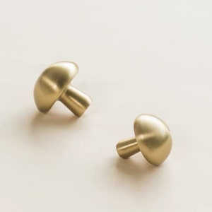 May include: Two gold mushroom-shaped cabinet knobs. The knobs have a smooth, rounded surface and a short stem.