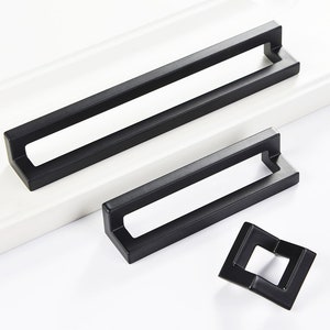 以下が含まれることがあります： 中央に切り抜きのある長方形の黒い金属製のキャビネットプルが3つあります。プルはサイズが異なります