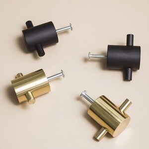 Könnte beinhalten: Vier Schrankknöpfe, zwei in Schwarz und zwei in Gold, mit einem T-förmigen Griff. Die Knöpfe sind zylindrisch und haben eine Schraube auf der Rückseite.