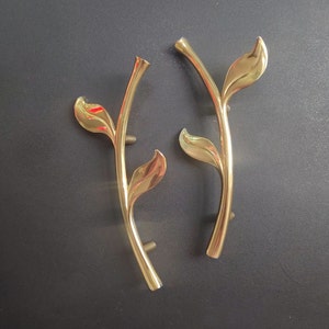 以下が含まれることがあります： 葉っぱが付いたスタイリッシュな枝の形をした、金色の金属製のキャビネットドアの取っ手2つ