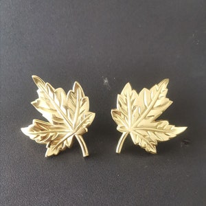 May include: A pair of gold-tone maple leaf earrings. The leaves are detailed and have a textured surface.