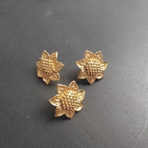 Op de afbeelding: Drie gouden zonnebloemvormige oorbellen. De oorbellen zijn gedetailleerd met een getextureerd midden en hebben een licht verhoogde rand.