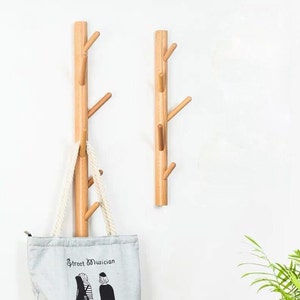 Pode incluir: Dois ganchos de parede de madeira em forma de galhos de árvore com múltiplos ganchos. Uma bolsa tote azul claro com o texto "Street Musician" e uma ilustração em preto e branco de duas pessoas está pendurada em um dos ganchos.