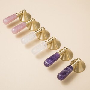 May include: Six cabinet knobs with a brass base and a crystal stone handle. The stones are pink, clear, and purple.