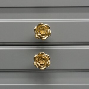 Può includere: Due tiretti per cassetti a forma di fiore dorati su una cassettiera grigia.