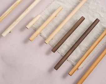 Op de afbeelding: Assortiment van houten kastgrepen in verschillende lengtes en afwerkingen, waaronder naturel hout en donker walnoot. De grepen zijn gerangschikt op een lichtroze oppervlak en een stuk beige linnen stof.