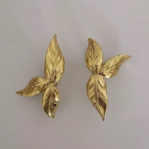 Op de afbeelding: Een paar gouden oorringen in de vorm van bladeren. Elke oorring heeft drie bladeren, waarbij het middelste blad het grootste is.