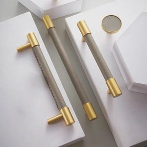 May include: Three gold cabinet pulls with gray leather wrapped handles. The pulls are shown on a white surface.
