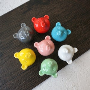 May include: Seven ceramic bear head knobs in various colours, including red, grey, blue, pink, yellow, white and green. The knobs are all approximately 5 cm in diameter and have a smooth, glossy finish.