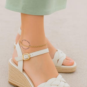May include: Gold-colored chain anklet with a delicate design. The anklet is worn on an ankle with a tattoo that reads "IN THE END, BEGIN AGAIN." The ankle is paired with white sandals with a woven espadrille wedge heel.