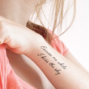 Op de afbeelding: Een vrouwenarm met een tijdelijke tatoeage met de tekst "Excuse me while I kiss the sky."