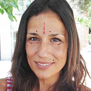 Puede incluir: Una mujer con el pelo largo y castaño lleva una camisa a rayas rojas, blancas y azules. Tiene un bindi rojo y dorado adornado con joyas en la frente y acentos rojos adornados con joyas en la cara.