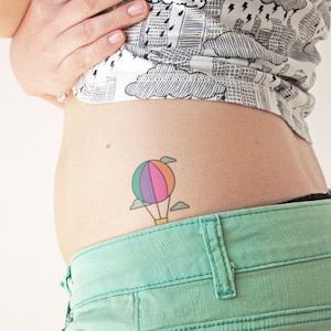 Puede incluir: Un tatuaje de globo aerostático colorido con nubes, una cesta y una cuerda amarilla. El tatuaje está en el abdomen inferior de una persona.