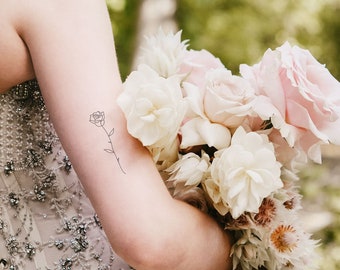 Könnte beinhalten: Eine Braut hält einen Strauß weißer und rosa Rosen. Ein einfaches Linienbild einer Rose ist auf ihrem Arm tätowiert.