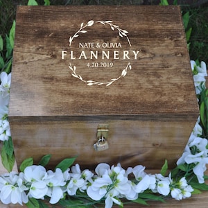 May include: A wooden box with a lock and key, decorated with a white floral wreath and the text "Nate & Olivia Flannery 4.20.2019".