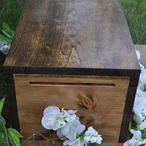 May include: A wooden box with a dark brown stain and a natural wood finish. The box has a hinged lid and a small slot on the front. The box is sitting on a wooden surface with white flowers.