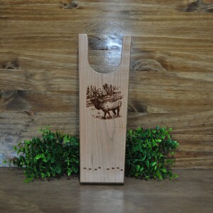 May include: Wooden boot jack with an engraved image of an elk and a hunter in a forest setting. The boot jack has a brown stain and a series of small holes near the bottom.