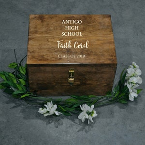 May include: A wooden box with a lock and key. The box is engraved with the text "Antigo High School" and "Faith Corel, Class of 2019". The box is surrounded by white flowers and green leaves.