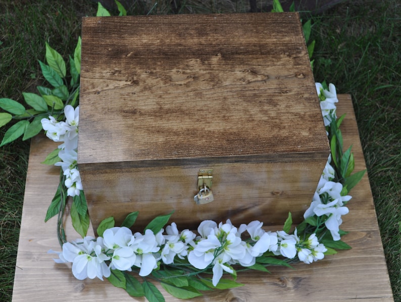 May include: A wooden box with a lock and key, decorated with a garland of white flowers and green leaves.