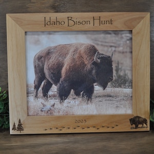 May include: A wooden picture frame with a photo of a bison in a field. The frame is engraved with the text "Idaho Bison Hunt 2023" and has a small bison silhouette in the bottom right corner.