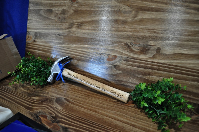 May include: A small wooden hammer with a silver head and a blue ribbon tied around the handle. The handle is engraved with the text "When it comes to being a good man, you've Nailed it!"