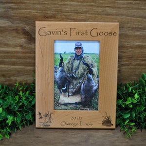 May include: A wooden photo frame with a personalized inscription that reads "Gavin's First Goose 2020 Oswego Illinois". The frame features a photo of a person holding two geese.