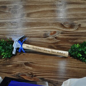 May include: A silver hammer with a wooden handle engraved with the text "When it comes to being a good man, you've Nailed it!" The hammer is tied with a blue ribbon and surrounded by green foliage.