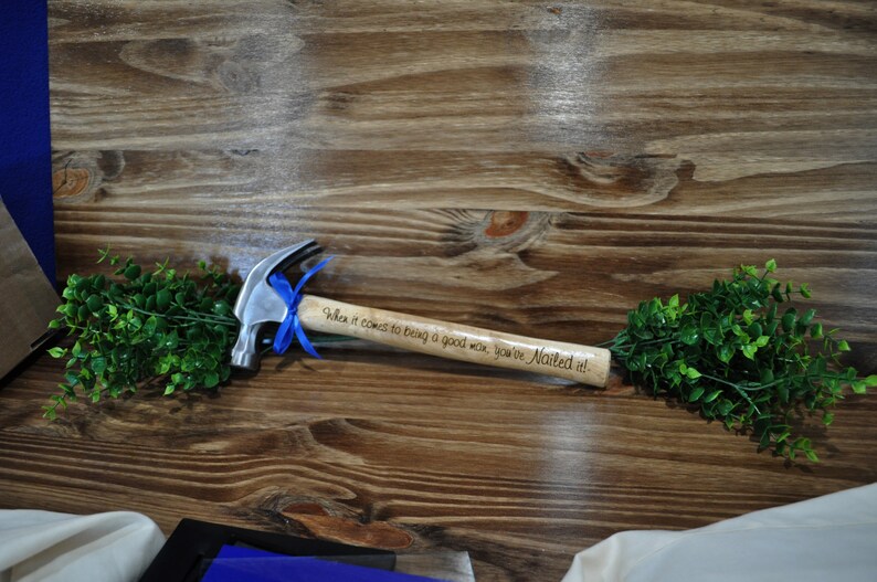 May include: A silver hammer with a wooden handle engraved with the text "When it comes to being a good man, you've Nailed it!" The hammer is tied with a blue ribbon and surrounded by green foliage.