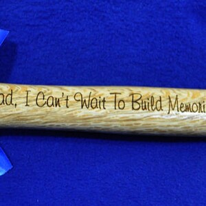 May include: A 16 oz hammer with a wooden handle. The handle is engraved with the text "Dad, I Can't Wait To Build Memories Together! Love, Cora". A blue ribbon is tied around the hammer head.