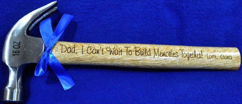 May include: A 16 oz hammer with a wooden handle. The handle is engraved with the text "Dad, I Can't Wait To Build Memories Together! Love, Cora". A blue ribbon is tied around the hammer head.