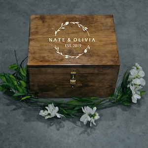 May include: A wooden box with a lock and a white inscription that reads "Nate & Olivia Est. 2019" surrounded by a wreath design. The box is sitting on a gray surface with green leaves and white flowers.
