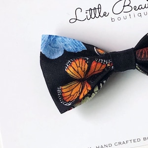May include: A black bow tie with a colourful butterfly design in orange, blue, and white. The bow tie is handmade and features the text "Little Beau boutique".