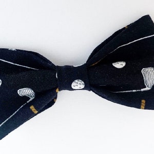 May include: A black bow tie with a golf theme. The fabric features a pattern of white golf balls, golf clubs, and gold accents. The bow tie is a classic accessory for formal or semi-formal attire.