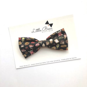 Könnte beinhalten: Eine schwarze Fliege mit einem sich wiederholenden Muster aus Cartoon-Essen, darunter Pizza, Eiscreme und Sushi. Die Fliege liegt auf einer weißen Karte mit dem Text "Little Beau boutique" und "HIGH QUALITY, HAND CRAFTED BOW TIES FOR INFANTS, TODDLERS & YOUTH."