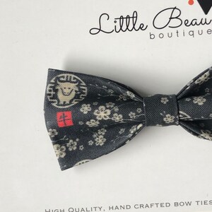 May include: A dark grey bow tie with a Japanese-inspired pattern. The design includes a bull emblem, cherry blossoms, and red characters. The bow tie is handmade and has a classic shape.