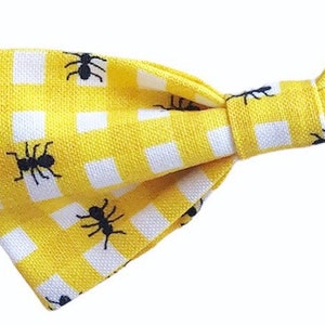 May include: A yellow bow tie featuring a white gingham pattern and black ant motifs. The fabric bow tie is tied in a classic shape. This accessory is suitable for formal or casual wear. The bow tie is made of fabric.