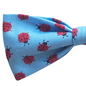 May include: A light blue bow tie featuring a repeating pattern of red ladybirds. The bow tie is made of fabric and is tied in a classic bow shape. The ladybirds have black spots and antennae.